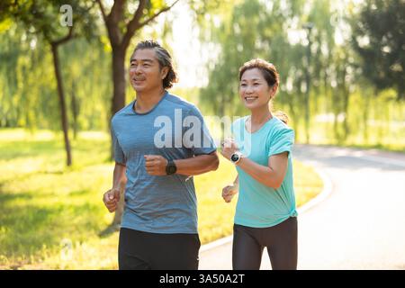 Joyeux couple chinois dans leurs 50 ans profiter d'une course de jour à travers un parc verdoyant de la ville. La paire porte des vêtements sportifs et une smartwatch, soulignant un mode de vie sain et la vitalité. Cette image est idéale pour les sujets sur le fitness, les activités de couple et les loisirs de plein air à Pékin. Banque D'Images