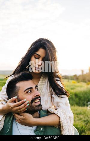 Jeune couple profite d'une journée d'été à la campagne ; un homme donne un tour de piggyback à sa petite amie sur une prairie verte. Moment de style de vie romantique dans un cadre naturel. Convient pour le voyage, l'extérieur et la marque relationnelle. Banque D'Images