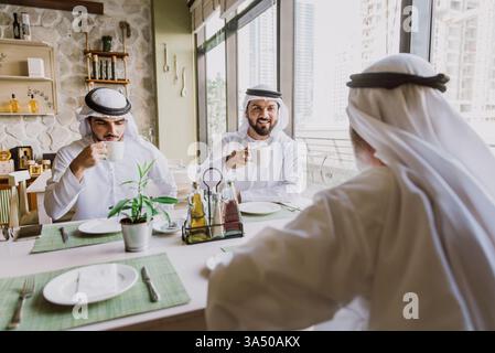 Trois hommes d'affaires partagent le thé dans un café de Dubaï, habillés de kanduras émiratis traditionnels. La scène allie l'étiquette des affaires à l'hospitalité du moyen-Orient dans un cadre culturel. Adapté aux thèmes d'entreprise, de voyage et d'affaires régionaux. Banque D'Images