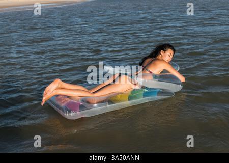 Femme Latina avec les yeux fermés se relaxant sur un matelas gonflable flottant dans la mer. Elle profite d'un moment d'été insouciant avec l'eau et la lumière du soleil. Idéal pour les vacances, les voyages et les campagnes de style de vie à la plage. Banque D'Images
