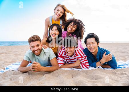 Scène lumineuse et insouciante d'amis divers se prélassant sur une plage de sable fin en été. Le groupe sourit alors qu'ils se détendent au bord de la mer, rayonnant d'amitié et de vibrations de vacances. Idéal pour les voyages, le style de vie et les campagnes multiculturelles célébrant les loisirs estivaux et la convivialité. Banque D'Images