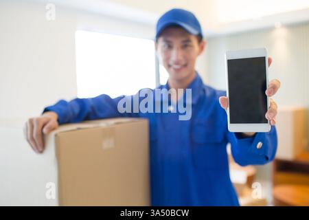 Un motard chinois souriant dans un uniforme bleu tient une boîte et montre un smartphone à l'intérieur d'un cadre de maison. Idéal pour le service de déménagement, la logistique et les concepts de déménagement avec une ambiance amicale et professionnelle. Scène intérieure claire adaptée pour illustrer le déménagement, la livraison et la connectivité des ménages. Banque D'Images