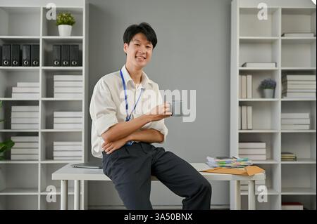 Un beau et heureux jeune homme d'affaires asiatique est assis à un bureau moderne, tenant une tasse de café. Cette image professionnelle convient aux campagnes de finance, de banque et de style de vie sur le lieu de travail. Banque D'Images