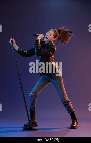 La chanteuse chinoise tient un micro et chante sur fond bleu de studio. Ce cliché de performance studio capture la musique contemporaine et la présence sur scène avec espace de copie pour le texte. Convient pour la musique, le divertissement et les fonctionnalités vocales pop. Banque D'Images