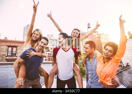 Femme noire souriante avec son bras autour d'un homme asiatique, rejoint par des amis sur un toit. Photo de style de vie urbain contemporain parfaite pour les amitiés, les célébrations et la vie urbaine. Le décor sur le toit offre une toile de fond élégante pour les campagnes sociales. Banque D'Images