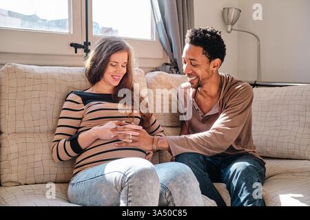 Portrait de style de vie chaleureux d'un couple multiethnique à la maison. Un homme noir touche doucement le ventre de sa femme enceinte alors qu'ils sont assis sur un canapé, souriant avec un bonheur attendu. Banque D'Images