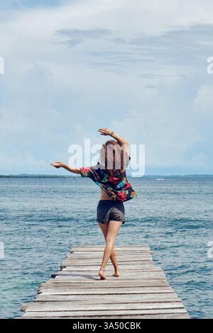 Femme hispanique aux cheveux bouclés lève les bras tout en se promenant le long d'une passerelle en bois à la plage dans la journée. La scène capture une ambiance estivale insouciante avec vue sur la mer et lumière naturelle. Parfait pour les voyages, les vacances et les campagnes de style de vie en plein air. Banque D'Images