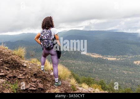 Vue arrière d'une randonneuse hispanique à un sommet rocheux, sans visage montré alors qu'elle regarde à travers un vaste paysage. Cette image de voyage capture la solitude et l'aventure en plein air. Idéal pour la randonnée, l'alpinisme et les campagnes de paysage de destination. Banque D'Images