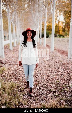 Femme hispanique souriante portant un chapeau marche le long d'un chemin bordé d'arbres dans un parc d'automne. La lumière Golden Hour met en valeur les feuilles d'automne et la tenue décontractée comme elle aime la saison. Une image chaleureuse et accessible adaptée aux campagnes de plein air, de voyage et de loisirs. Banque D'Images