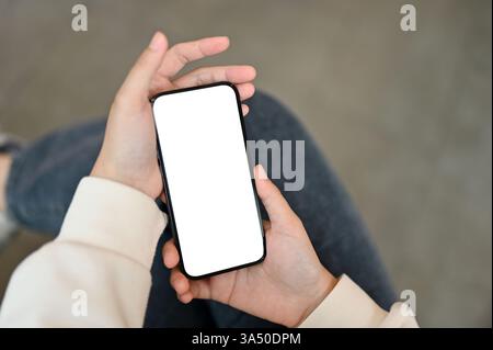 Vue de dessus d'une femme assise dans un café, tenant un smartphone avec une maquette d'écran blanc sur ses genoux. Cette image est idéale pour présenter des applications mobiles, les médias sociaux et la messagerie en ligne dans un cadre intérieur décontracté. Utilisez-le pour illustrer la connectivité et la technologie mobile dans des contextes de café ou de style de vie. Banque D'Images