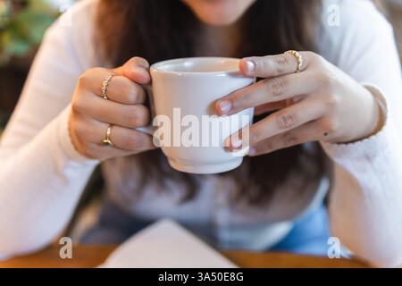 Femme hispanique assise dans un café tenant une tasse de café. Une image de style de vie détendue parfaite pour la culture du café, les réunions informelles et les moments de pause urbaine. Convient pour les blogs Lifestyle, le marketing de café et le contenu de résilience. Banque D'Images