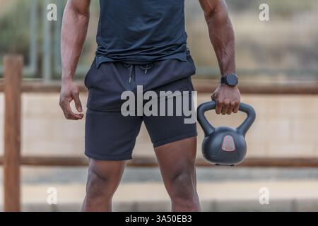 Homme noir en vêtements d'activité tenant une kettlebell dans un parc pendant la journée. Idéal pour le fitness, la musculation, les entraînements en plein air et les campagnes de style de vie sain. Une image polyvalente pour les contenus sportifs et de bien-être. Banque D'Images