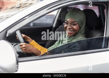 Joyeuse jeune femme musulmane noire portant un foulard sourit tout en conduisant sa nouvelle voiture. Elle est assise sur le siège avant, transmettant la propriété et l'excitation. Idéal pour les images automobiles, de style de vie et de voyage. Banque D'Images