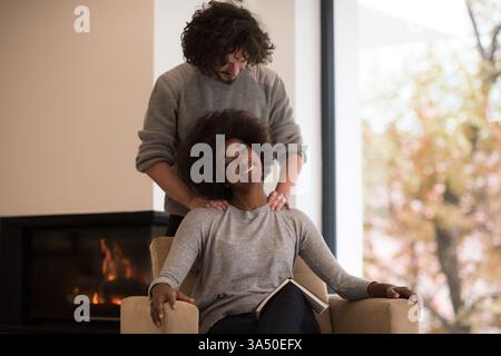 Jeune couple multiethnique embrasse par une cheminée confortable à la maison, lisant un livre pendant l'automne. La scène chaleureuse et intime fonctionne bien pour le style de vie à la maison, la romance et les campagnes saisonnières. Banque D'Images
