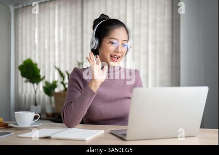 Femme asiatique souriante portant des lunettes et des écouteurs ayant un appel vidéo sur ordinateur portable tout en étant assis à table avec ordinateur portable et café à la maison dans la lumière du jour Banque D'Images