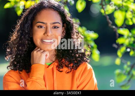 Une jeune femme afro-américaine souriante se tient à l'extérieur parmi les feuilles vertes, projetant une pose confiante et amicale. Un portrait frais adapté aux campagnes lifestyle, mode et outdoor. Banque D'Images