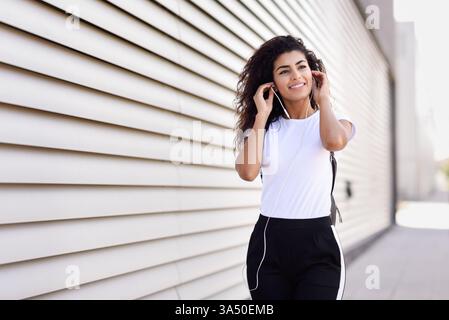 Une femme arabe joyeuse aux cheveux bouclés porte des écouteurs et se promène dans une rue urbaine ensoleillée. Un moment de style de vie animé adapté au contenu sur le style de la rue, la musique et la vie urbaine contemporaine. Idéal pour le style de vie, les voyages et les thèmes d'écoute mobile. Banque D'Images