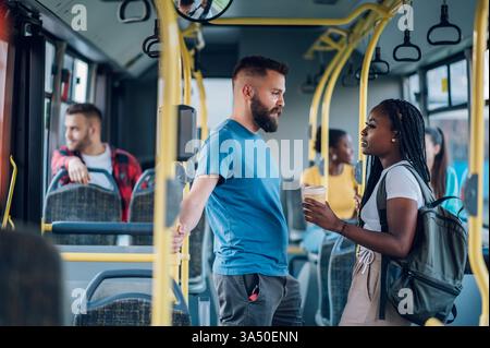 Groupe multiracial d'amis parlant et riant tout en se tenant debout dans un bus de la ville et en profitant du café. Ce moment diversifié et sociable convient au style de vie, aux voyages, aux trajets quotidiens et aux activités de groupe. Parfait pour les campagnes mettant en avant l'amitié et les expériences urbaines. Banque D'Images