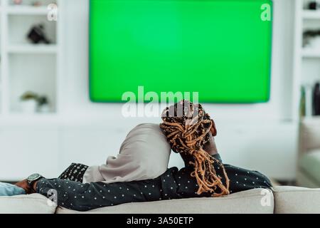 Un couple africain se détend à la maison sur un canapé, regardant la télévision avec un fond d'écran vert chroma. La femme porte un hijab, mettant en valeur la mode musulmane contemporaine dans un décor de salon. Idéal pour le style de vie, le divertissement à domicile et les scènes de famille. Banque D'Images