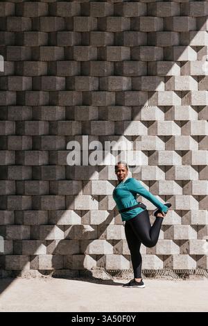 Femme afro-latina dans les écouteurs faisant l'exercice d'étirement debout contre le mur de briques à l'extérieur pendant l'entraînement le jour ensoleillé Banque D'Images
