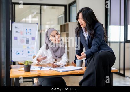 Amicale femme d'affaires musulmane asiatique dans le bureau partageant une conversation avec un collègue. Idéal pour le coworking, le travail d'équipe et le contenu diversifié du lieu de travail. Banque D'Images