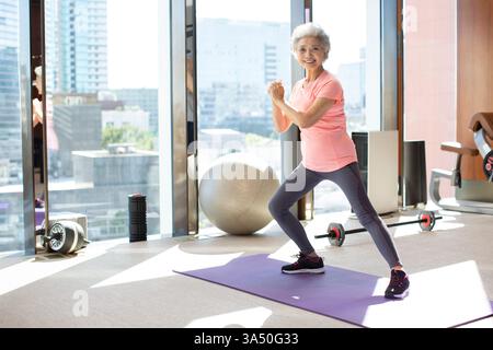 Femme chinoise âgée souriante effectuant un étirement debout dans une salle de sport lumineuse, mettant en valeur la vitalité et le bien-être. Convient pour le fitness senior, le vieillissement actif et les campagnes de mode de vie sain, avec un accent sur le bien-être. Banque D'Images