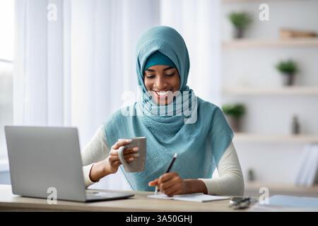 Portrait d'une femme musulmane souriante freelance travaillant à la maison dans un bureau à domicile lumineux. Elle verse du café, tape sur un ordinateur portable et écrit des notes dans un bloc-notes, avec un espace de copie sur le bureau. Idéal pour les sujets sur le travail à distance, les femmes entrepreneurs et le mode de vie professionnel. Banque D'Images