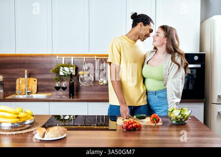 Un homme afro-américain souriant et une copine blanche Curvy partagent un moment chaleureux dans une cuisine moderne. Ils se regardent tout en cuisinant, exprimant la romance quotidienne et la vie domestique confortable. Cette scène est idéale pour le style de vie, les relations multiculturelles et les thèmes de la cuisine à la maison. Banque D'Images