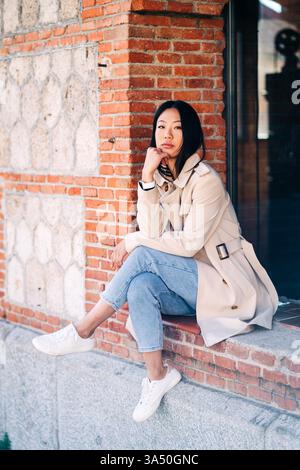 Femme asiatique assise avec les jambes croisées sur le mur de briques à l'extérieur Banque D'Images