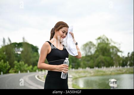 Femme asiatique fatiguée et en sueur dans les vêtements de sport prend une pause après cardio dans un parc public. Elle essuie la transpiration avec une serviette, capturant la forme physique en plein air et la récupération. Idéal pour les thèmes fitness, course et bien-être. Banque D'Images