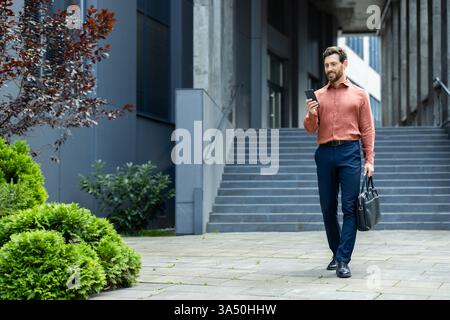 Homme d'affaires prospère marchant à travers la ville avec le téléphone dans les mains et le sac d'affaires. Homme en chemise tapant un message texte et naviguant sur les réseaux sociaux en pleine croissance. Banque D'Images