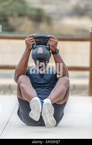 Homme noir déterminé dans les vêtements d'action est assis sur le sol dans un parc ensoleillé, faisant des abdos avec une kettlebell. Un portrait d'entraînement dynamique en plein air parfait pour les campagnes de fitness, de musculation et de vie saine. Idéal pour les vêtements de sport, les promotions de gymnastique et les messages de bien-être. Banque D'Images