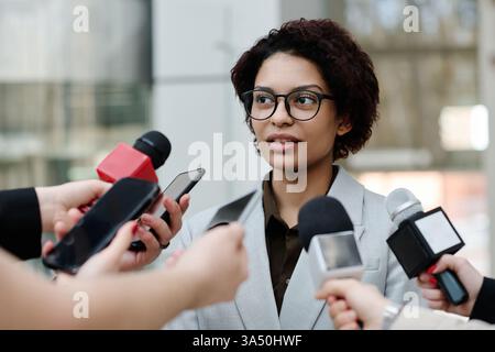Jeune femme d'affaires noire dans des lunettes donnant une interview à une conférence, parlant aux journalistes. Le cadre professionnel met en avant le leadership, la communication et l'engagement des médias. Idéal pour les affaires, les événements d'entreprise et les visuels d'entrevue. Banque D'Images