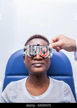 Femme noire subissant un examen de la vue portant une monture d'essai dans une clinique. Ce portrait de la santé met en évidence l'optométrie, les soins oculaires et l'examen professionnel. Une image claire et informative adaptée aux sujets médicaux et visuels. Banque D'Images