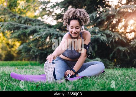 Scène de fitness dynamique en plein air avec une femme hispanique avec des écouteurs effectuant un virage assis vers l'avant dans un parc d'été. Image de style de vie lumineuse et énergique adaptée aux campagnes de bien-être, de remise en forme et d'exercice. Banque D'Images