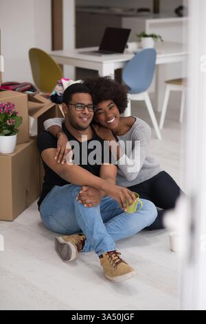 Joyeux couple afro-américain se relaxant dans leur nouvelle maison, assis sur le sol avec du café tandis que des boîtes mobiles les entourent. Une scène chaleureuse et relationnelle idéale pour la relocalisation, la vie à domicile et les campagnes de style de vie familial. Banque D'Images