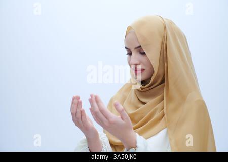 Élégante femme musulmane portant le hijab effectue un geste de prière traditionnel avec les mains levées dans la supplication. Isolé sur fond blanc, ce portrait respectueux capture la dévotion spirituelle et la culture islamique. Convient pour les campagnes fondées sur la foi, les caractéristiques culturelles ou le matériel d'éducation religieuse. Banque D'Images