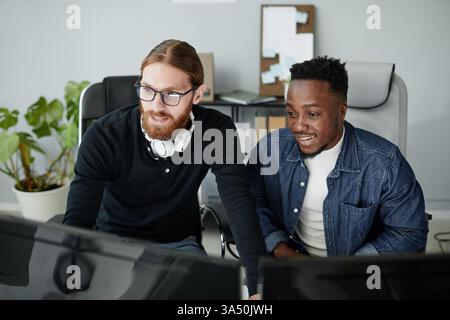 Groupe joyeux et diversifié de jeunes programmeurs travaillant ensemble dans un bureau moderne. Ils se concentrent sur les moniteurs d'ordinateur, mettant l'accent sur le travail d'équipe, la collaboration et le développement de logiciels. Cette image convient aux campagnes technologiques d'entreprise, au recrutement et au message de diversité. Banque D'Images