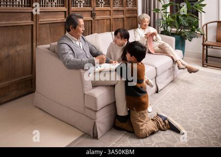 Grand-père chinois partageant une histoire avec ses petits-enfants dans un salon confortable. Ils se rassemblent autour d'un canapé avec un livre, profitant de la vie domestique et des liens familiaux. Idéal pour le style de vie, le rôle parental et les moments intergénérationnels. Banque D'Images