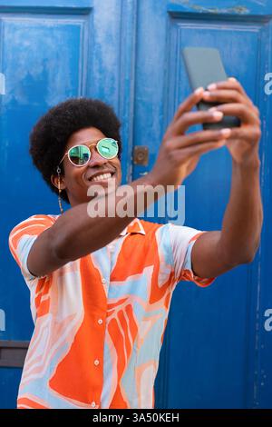 Homme afro-américain souriant avec un afro prend un selfie sur un smartphone sur un fond de rue bleu. Ce portrait extérieur optimiste convient parfaitement aux thèmes de mode de vie, de médias sociaux et de mode urbaine. Idéal pour l'image de marque personnelle et les visuels de style urbain contemporain. Banque D'Images
