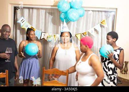 Joyeuse famille noire de cinq célébrant un anniversaire à la maison. Ils se tiennent parmi les décorations de fête colorées, un gâteau et des ballons, capturant la chaleur et l'amour de la famille. Une scène intérieure parfaite pour le style de vie familial, la célébration et les histoires de convivialité. Banque D'Images