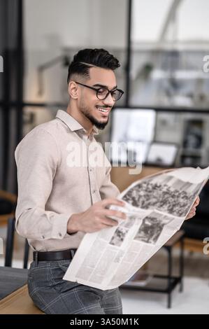 Joyeux jeune homme barbu dans des verres regarde un journal à l'intérieur, debout et appuyé sur une table pendant la journée. La scène transmet des informations et une ambiance professionnelle confiante et élégante. Idéal pour les affaires, les actualités et le contenu de style de vie des travailleurs indépendants. Banque D'Images