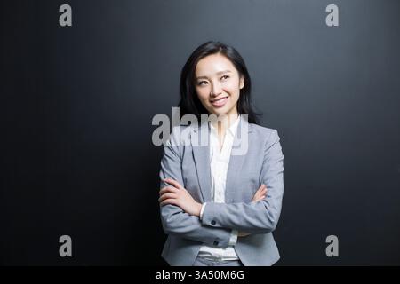 Femme d'affaires chinoise en costume se tient dans un studio, profondément dans la pensée avec une expression contemplative. Cette image de face sur fond noir traduit le professionnalisme et la concentration pour les campagnes d'entreprise, de leadership ou axées sur les idées. Banque D'Images