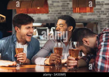 Groupe diversifié d'hommes se relaxant dans un bar, buvant de la bière et partageant la conversation. La scène décontractée convient au contenu sur l'amitié, la vie nocturne et la dynamique de groupe. Des verres lager visibles et une atmosphère joyeuse transmettent des moments sociables. Banque D'Images