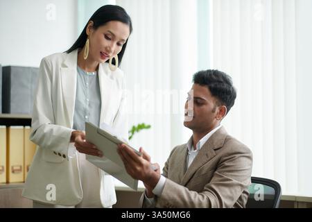 Une femme d'affaires asiatique souriante présente un rapport à un collègue indien masculin à travers une table près d'une fenêtre. L'environnement de bureau met l'accent sur le travail d'équipe et la communication professionnelle. Idéal pour les réunions d'affaires, la collaboration interculturelle et les environnements d'entreprise. Banque D'Images
