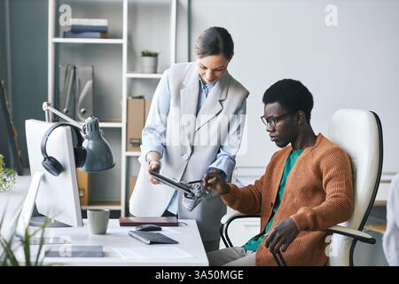 Femme d'affaires souriante avec un bras prothétique présentant un document sur une tablette à un collègue. Cette image met en évidence l'inclusion des personnes handicapées, le travail d'équipe et la technologie de bureau moderne. Adapté aux communications d'entreprise et aux campagnes inclusives sur le lieu de travail. Banque D'Images