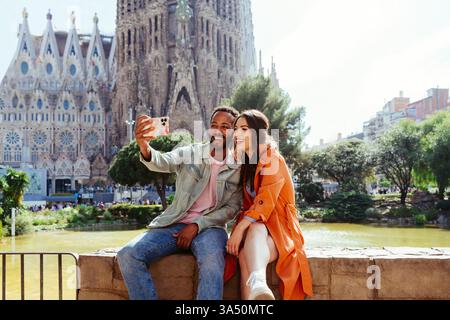Couple multiracial profitant de vacances à Barcelone près de la Sagrada Familia, capturant un selfie lors de leur visite de la ville. Cette scène d'amour de voyage met en valeur la romance, le tourisme urbain et la compagnie multiculturelle. Banque D'Images