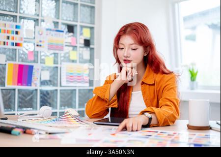 Graphiste féminin asiatique avec des cheveux bouclés rouges travaille à son bureau dans le studio, sélectionnant des couleurs à partir d'échantillons de couleur. Environnement de design moderne mettant en valeur la créativité et la sélection de couleurs. Banque D'Images