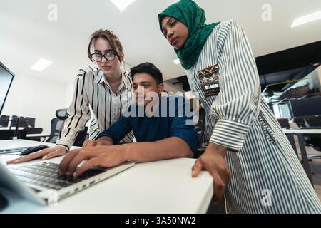 Équipe de démarrage multiethnique dans un bureau moderne ouvert. Une femme musulmane portant un hijab se joint à une séance de brainstorming tout en travaillant sur un ordinateur portable. La scène transmet la collaboration et le leadership diversifié, idéal pour les visuels de démarrage et de travail d'équipe. Banque D'Images