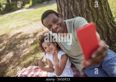 Un homme souriant à la peau foncée prend un selfie avec sa jeune fille dans un parc. Un moment familial chaleureux idéal pour les campagnes parentales, paternelles et lifestyle. Banque D'Images